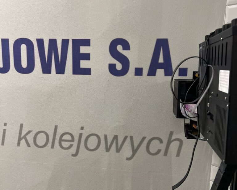 Zbliżenie liter napisu firmowego wydrukowanego UV na ścianie dworca kolejowego, obok głowica drukarki ściennej