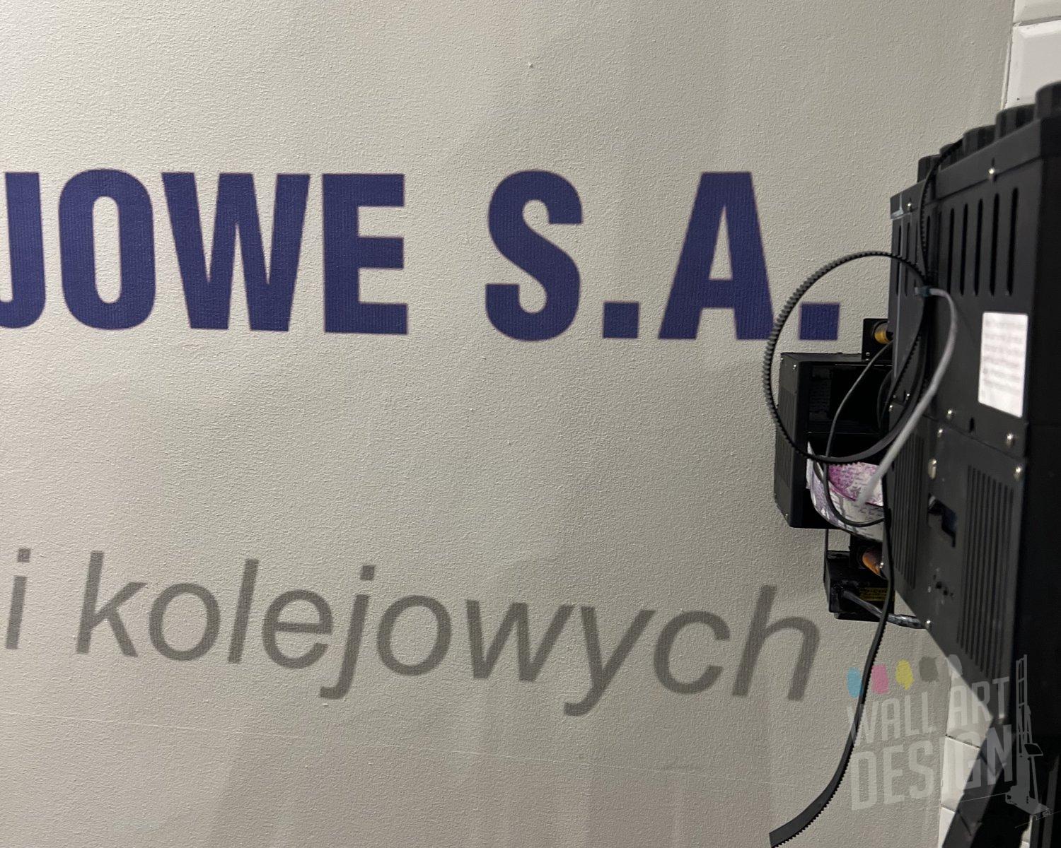 Zbliżenie liter napisu firmowego PKP PLK wydrukowanego UV na ścianie dworca kolejowego, obok głowica drukarki ściennej