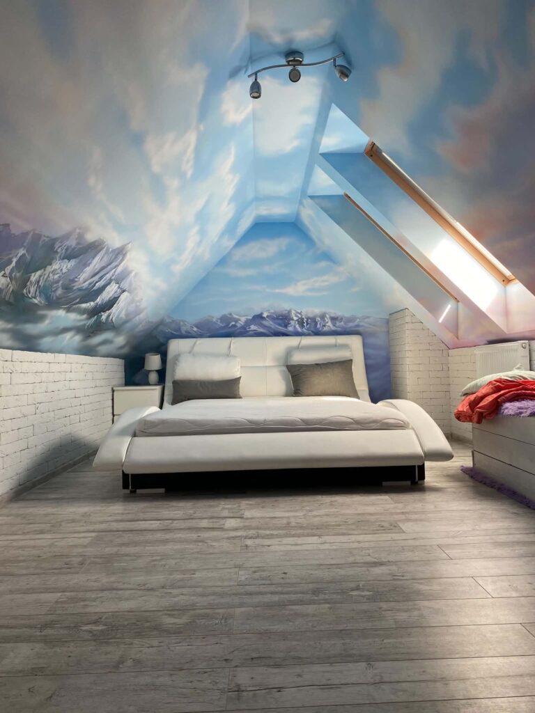 Mural malowany ręcznie w sypialni na poddaszu – niebo z chmurami i ośnieżone góry nad łóżkiem